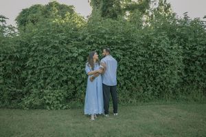Couple holding hands in front of lush green ivy wall outdoors, woman in blue dress, man in light shirt.