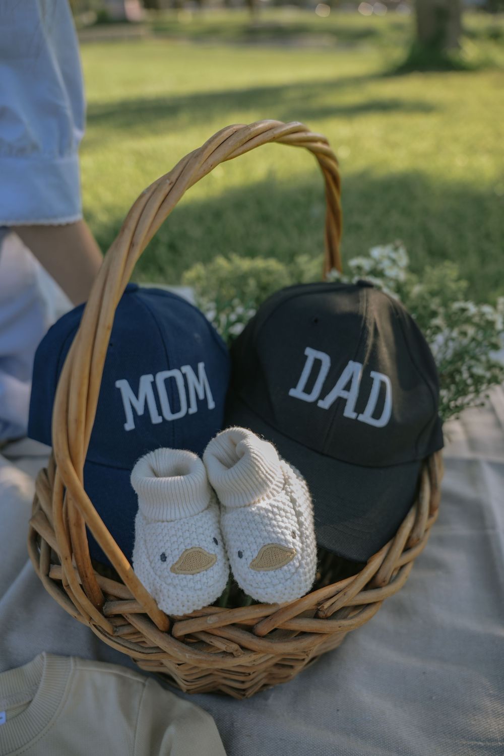 Wicker basket with navy Mom and dark Dad baseball caps and white knit baby booties on a picnic blanket outdoors.