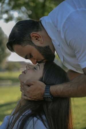 A bearded man in a white shirt tenderly kisses a woman's forehead while cradling her face outdoors.