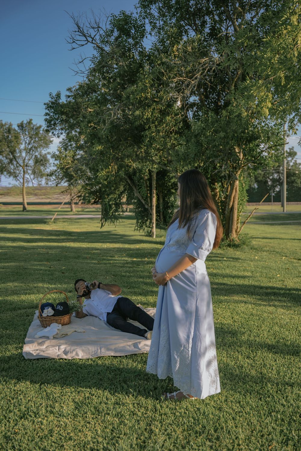 Pregnant woman in white dress stands in park while partner relaxes on blanket beneath green trees on sunny day.