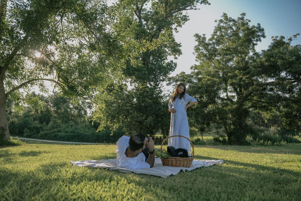 Two women enjoying a sunny picnic on a white blanket under large green trees in a lush park.