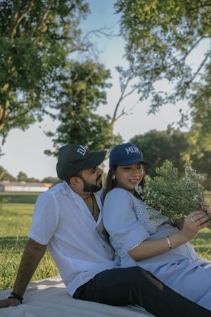 Couple sitting together on a blanket in a park, wearing matching navy caps, holding wildflowers on a sunny day.