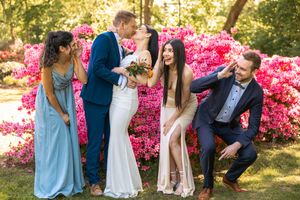 Bride and groom kissing surrounded by bridesmaids and groomsmen with vibrant pink azalea flowers in background.