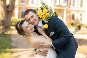 Happy newlyweds embracing outdoors, bride in white gown with yellow rose bouquet, groom in navy suit with boutonniere.