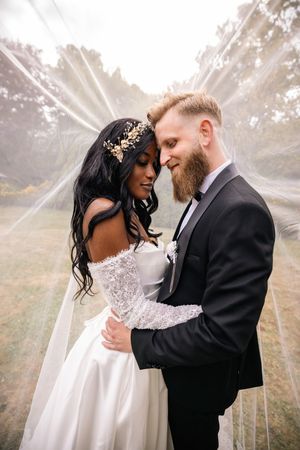 Interracial couple sharing romantic moment under wedding veil, bride in lace gown with gold crown, groom in black suit.