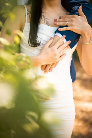 Couple embracing outdoors, woman in white dress with tattoo, both wearing rings, surrounded by greenery.