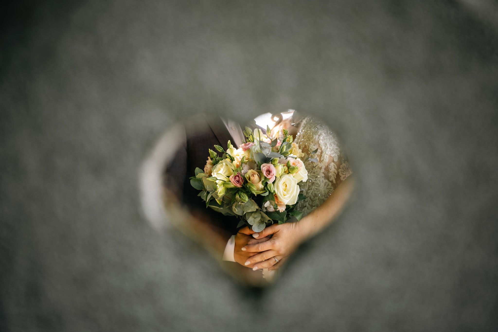 Hands holding a floral wedding bouquet with roses and hydrangeas, viewed through a heart-shaped opening.