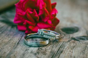 Two silver wedding rings with diamonds rest on rustic wood beside a vibrant red rose bloom.