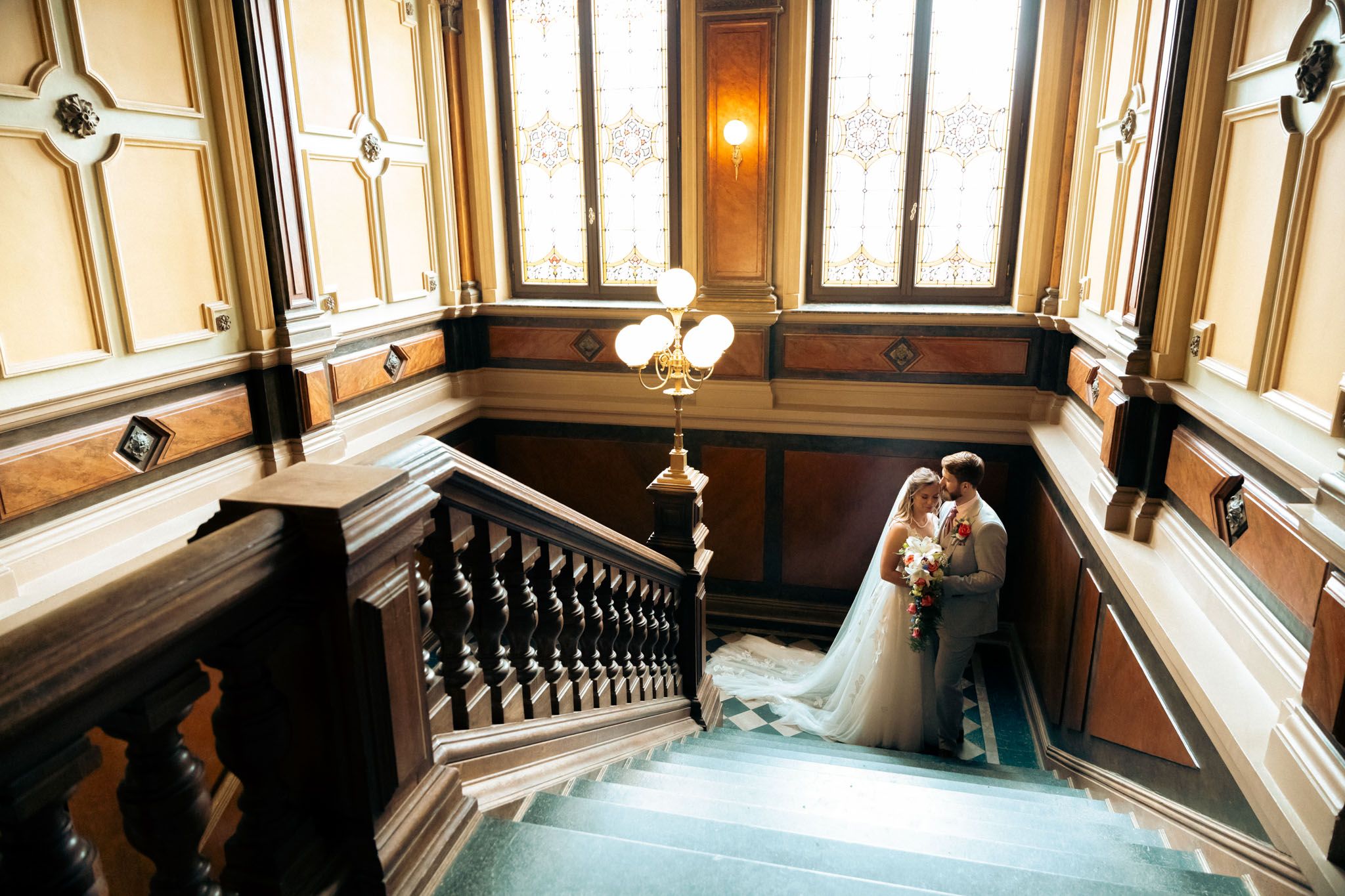 Bride and groom share a romantic embrace on an elegant grand staircase in a historic ornate venue.