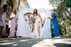 Bride and groom walk through a decorative banner held by wedding guests outdoors on a sunny day.