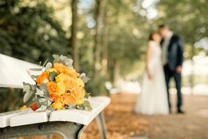 Orange rose wedding bouquet on white bench with blurred bride and groom kissing in bokeh background.