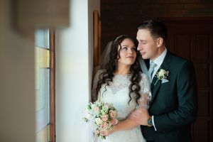 Elegant bride and groom posing by a window, bride holding a bouquet of pink and white flowers on their wedding day.