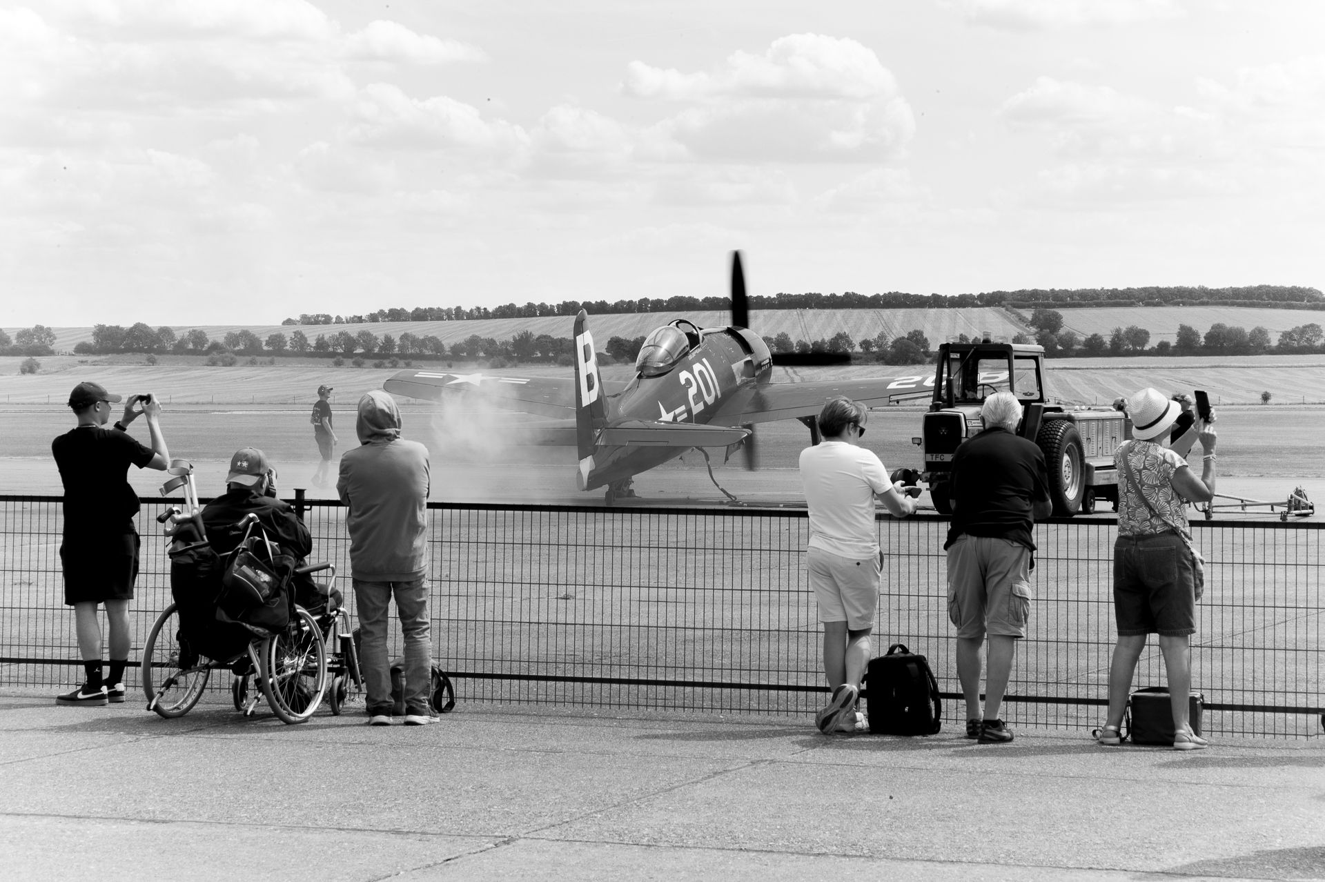 Black and white photo of spectators photographing a vintage WWII propeller aircraft at an airshow behind a fence.