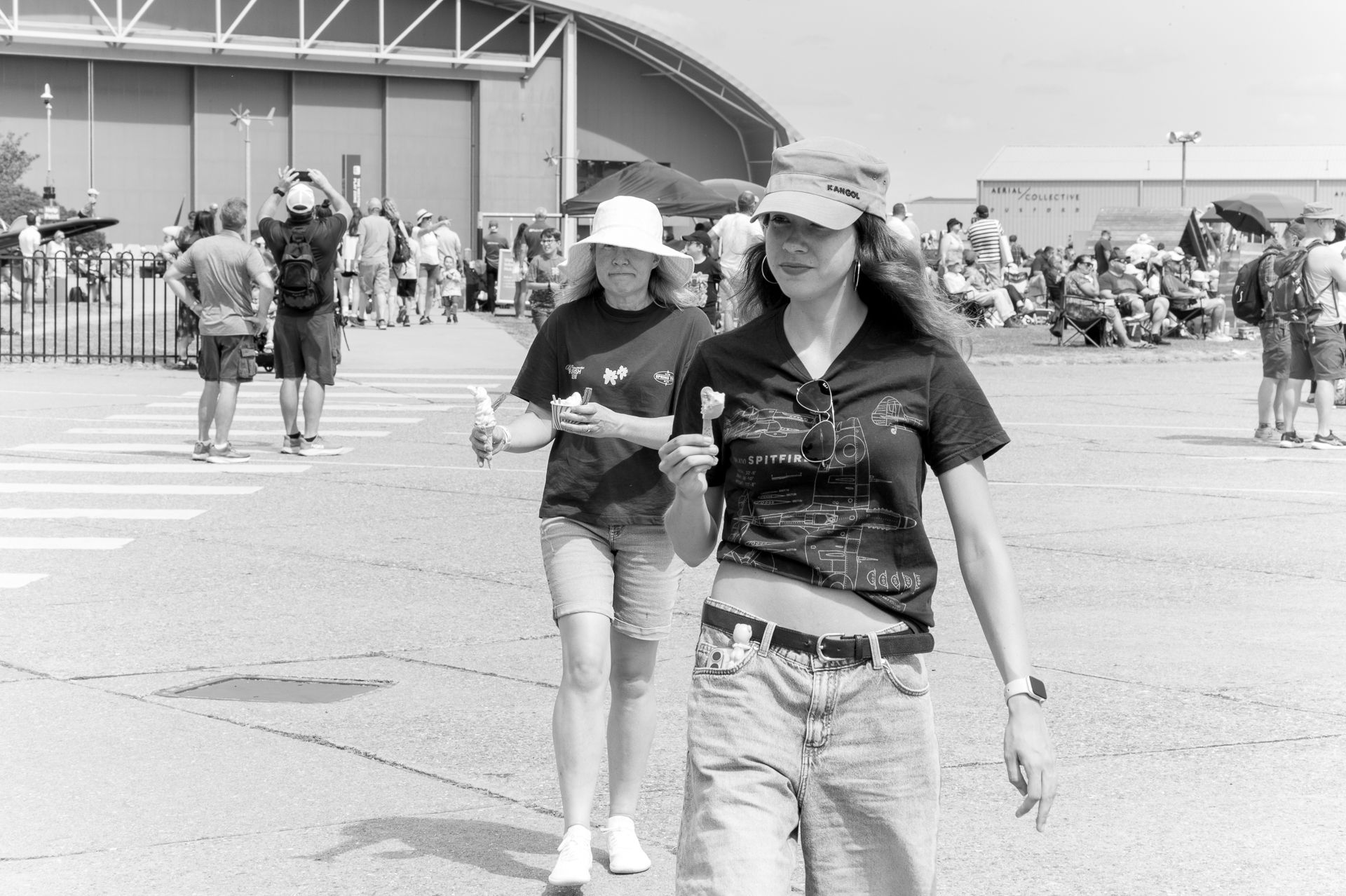 Two women wearing hats walk outdoors at a sunny event, with a crowd and large building visible in the background.