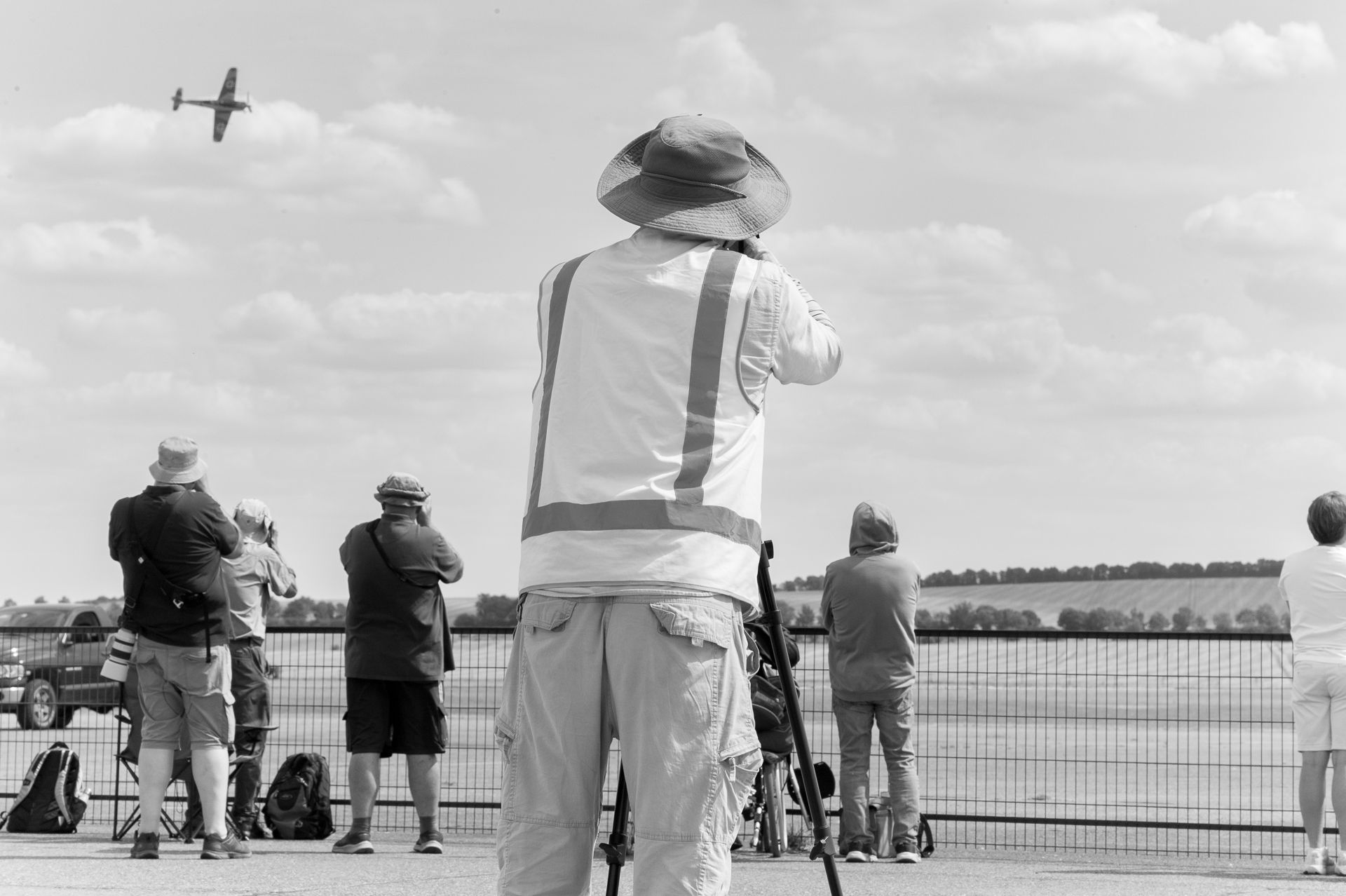 Black and white photo of spectators watching a vintage aircraft fly at an outdoor airshow event.