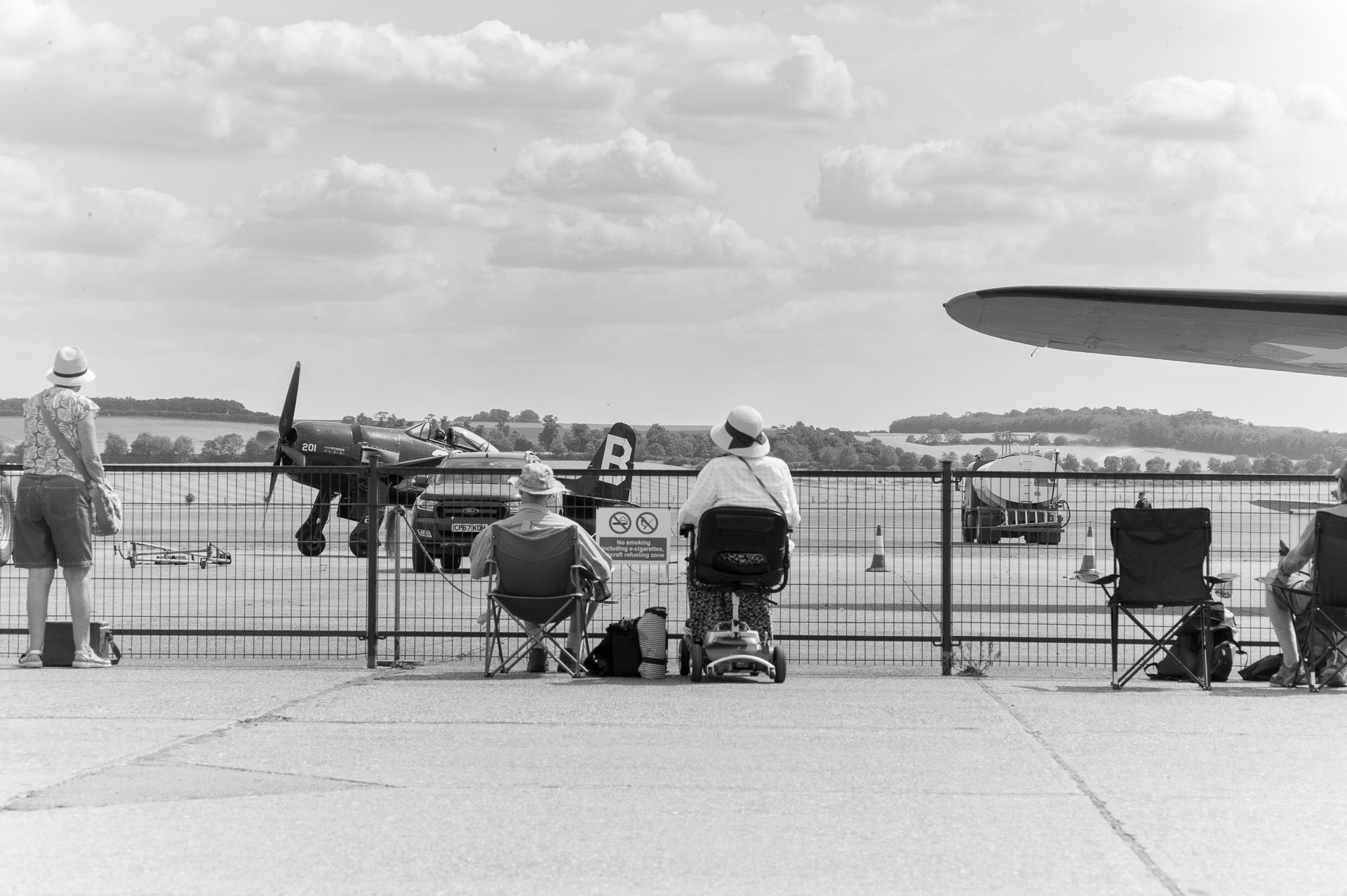 Black and white photo of spectators watching a vintage WWII propeller aircraft at an outdoor airshow.