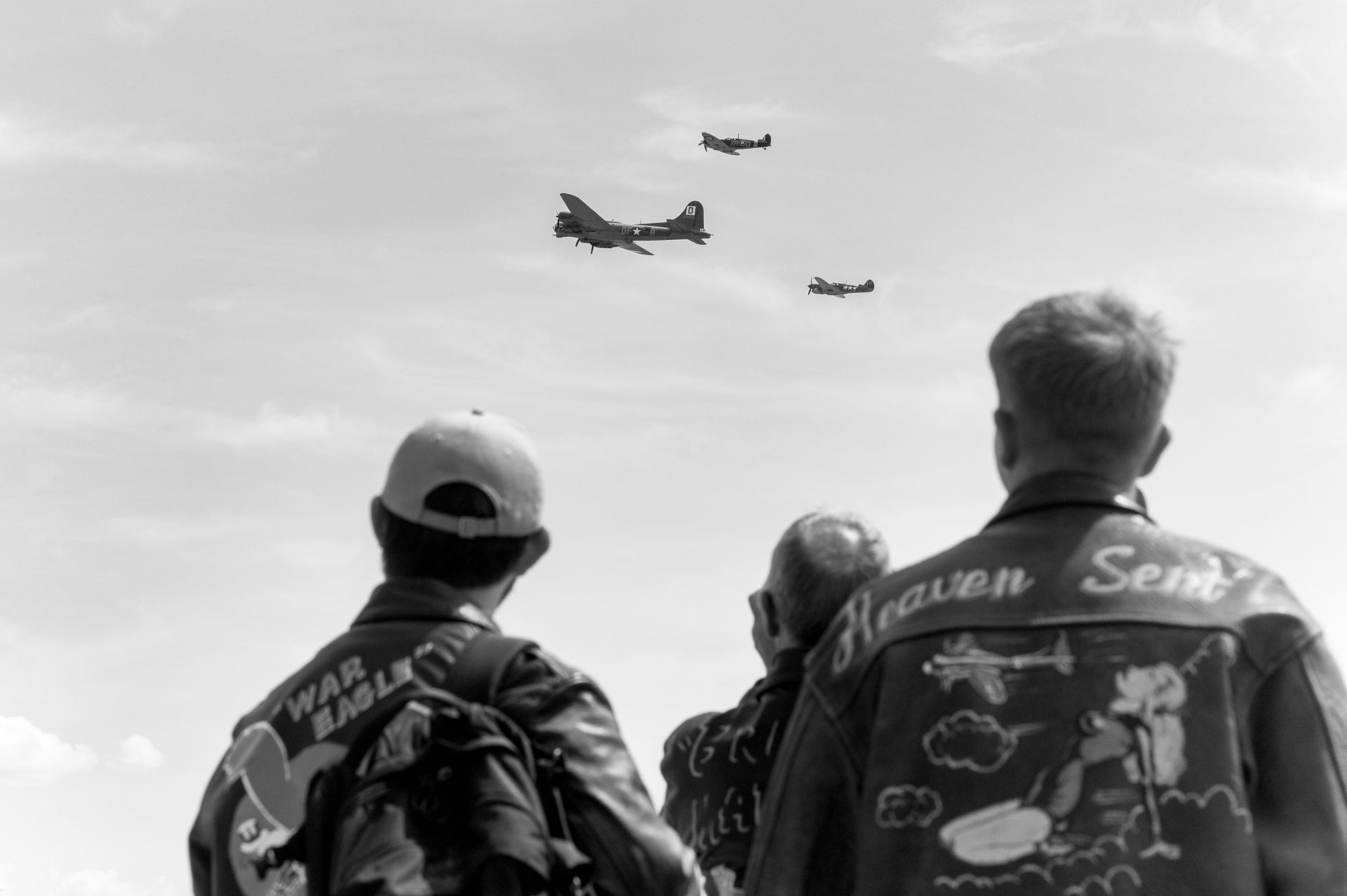 WWII veterans in leather jackets watch vintage B-17 bombers fly overhead at an airshow commemoration.