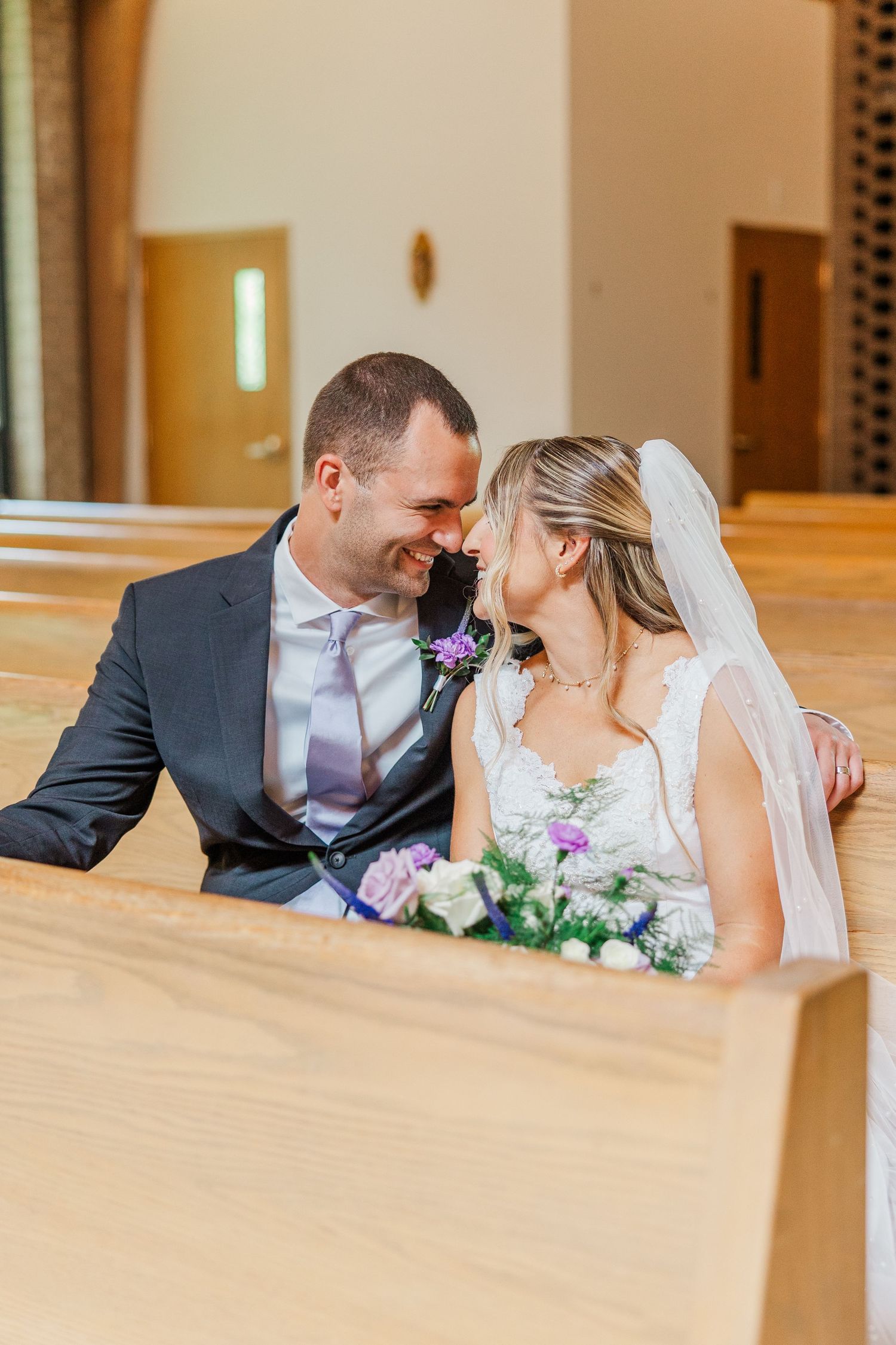 A bride and groom share an intimate moment on a church pew, with the bride holding a purple and white bouquet.
