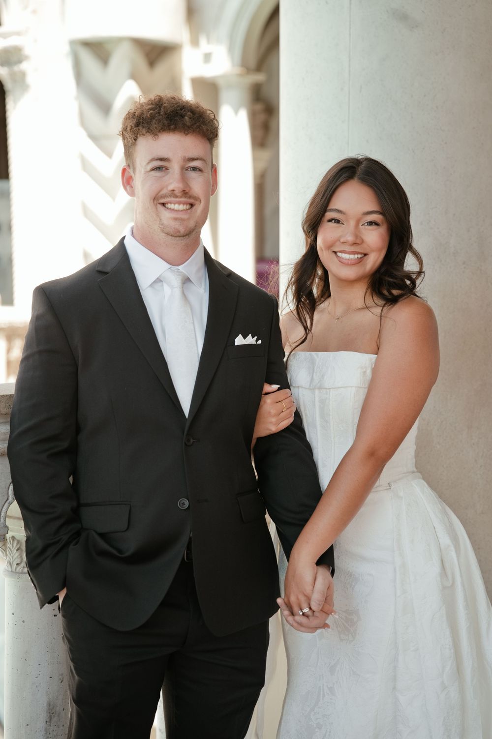 A smiling couple in wedding attire poses near stone columns, groom in black suit and bride in white strapless gown.