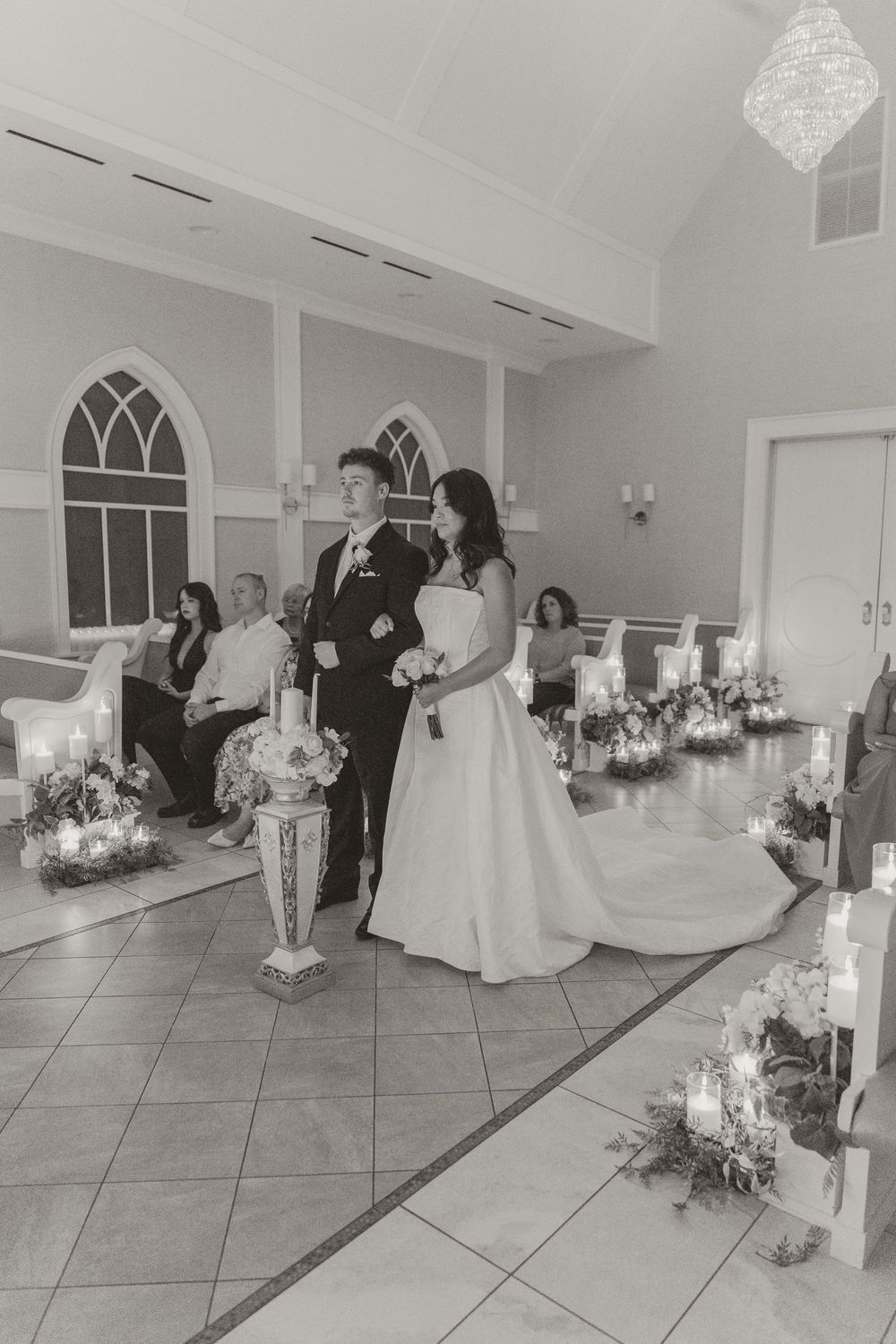 Black and white photo of bride and groom walking down the aisle in a chapel decorated with candles and floral arrangements.