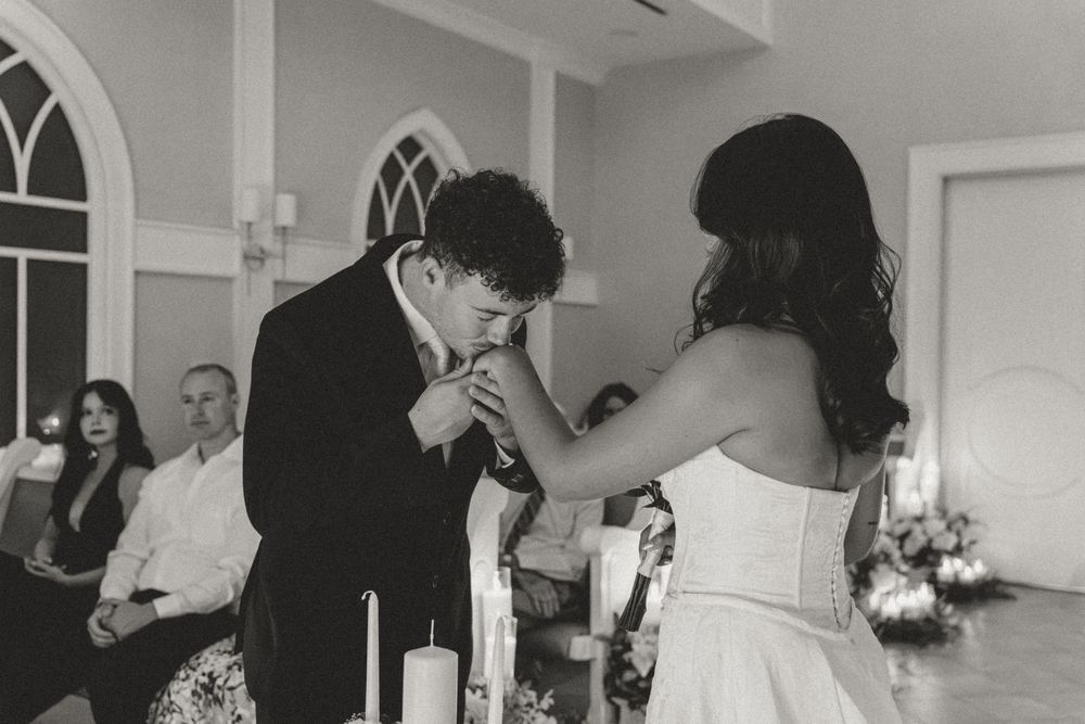 Groom kissing bride's hand at wedding ceremony altar with candles and guests in black and white photo.