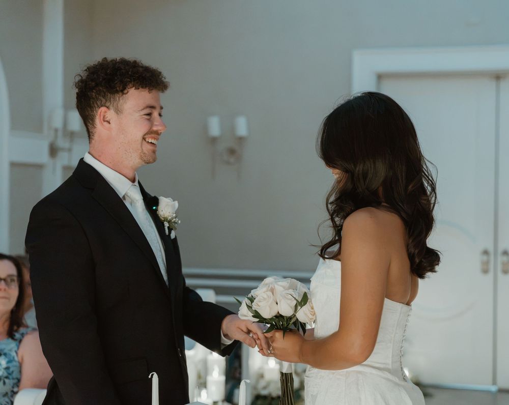 Smiling groom holds brides hands during wedding ceremony, bride holds white floral bouquet in elegant white dress.