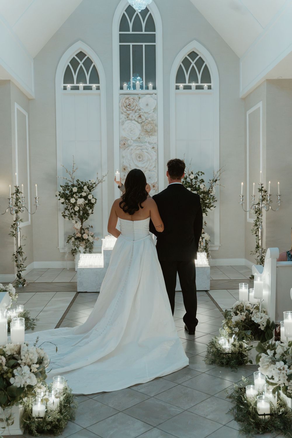 Bride and groom standing at chapel altar surrounded by candles and floral arrangements in elegant white wedding venue.
