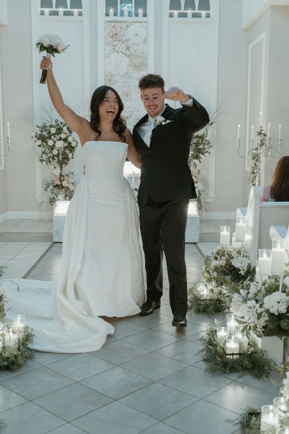 Joyful newlyweds celebrate after wedding ceremony, bride in white gown raising bouquet, groom in black suit, candles and florals.