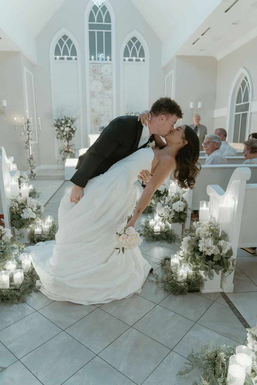 Bride and groom share a romantic dip kiss at the altar of a white chapel adorned with candles and floral arrangements.