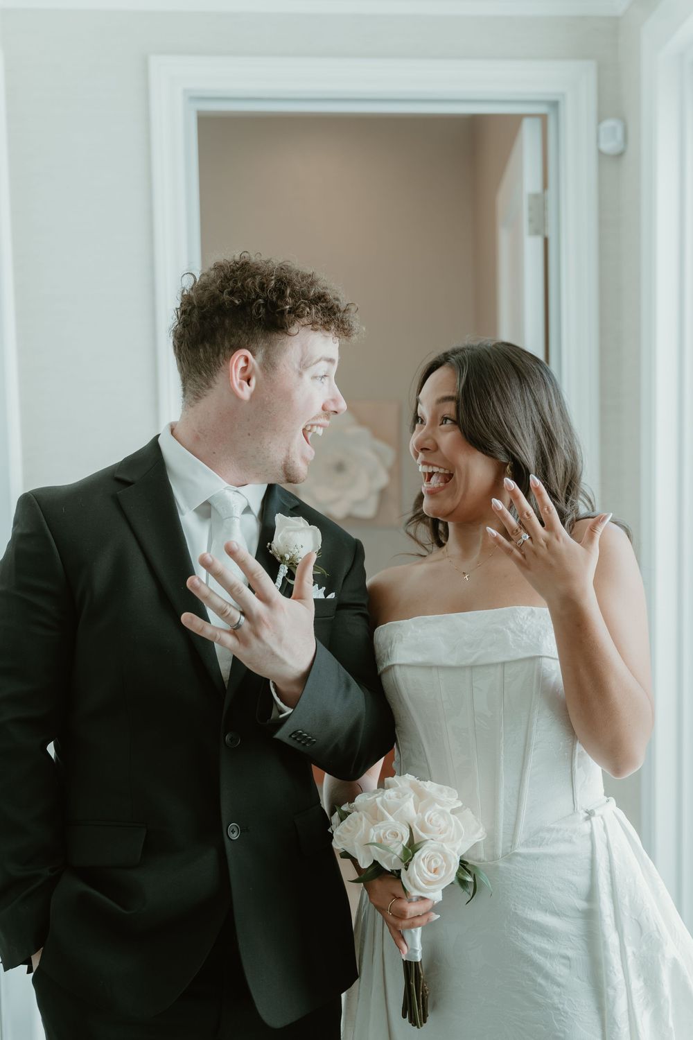 Newlywed couple in wedding attire joyfully showing off their wedding rings, bride holding white roses bouquet.