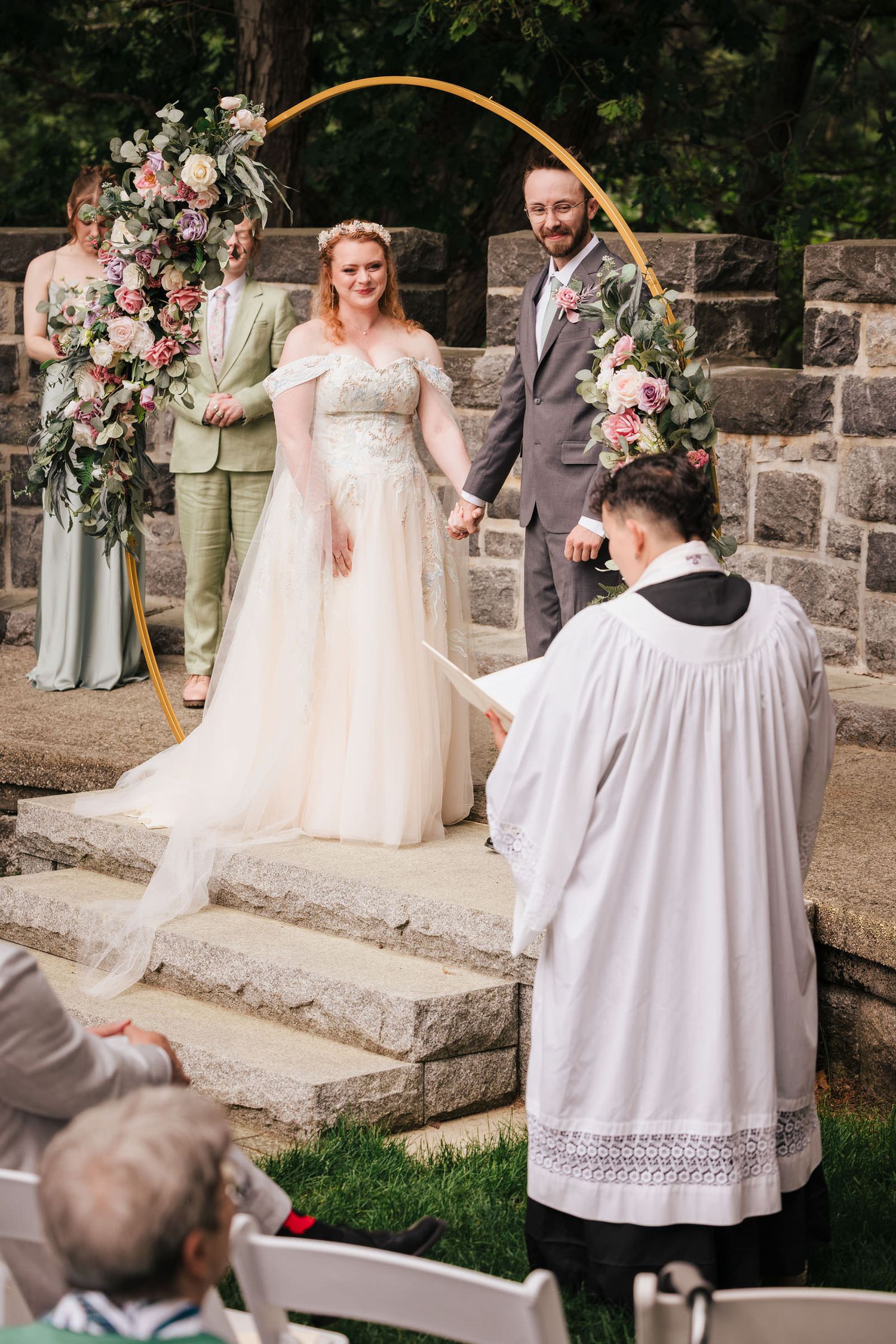 Reverend Claire officiating the wedding ceremony, bride and groom holding hands under a gold arch, wedding party looking on.