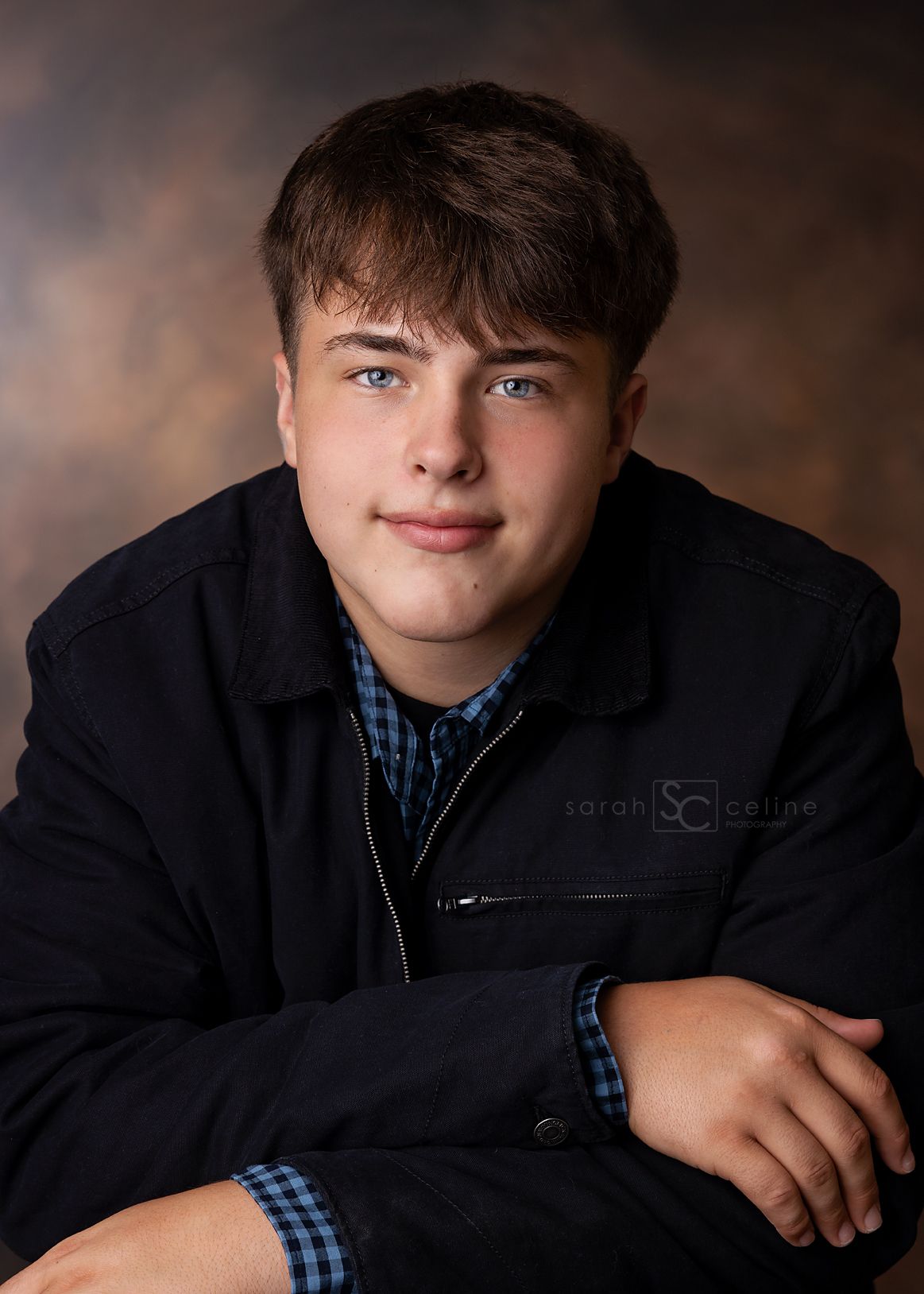A young man in a dark jacket and plaid shirt poses for a professional senior portrait against a studio backdrop.