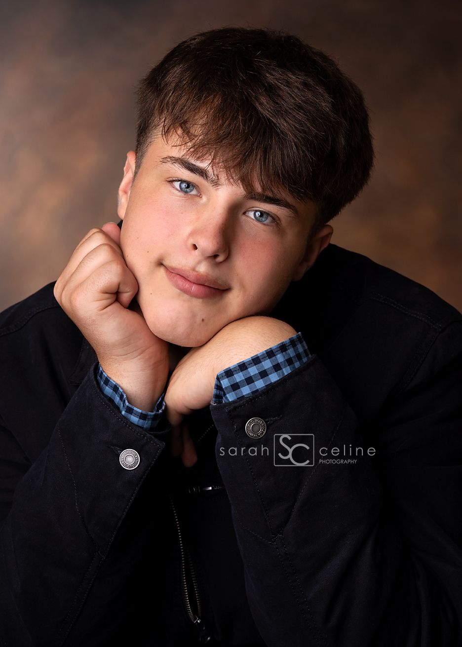 A teenage boy with blue eyes poses with hands under chin, wearing a black jacket over a blue plaid shirt, studio portrait.