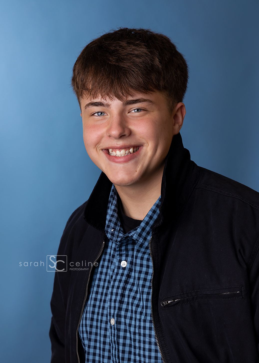 Smiling teenage boy in blue plaid shirt and black jacket poses for senior portrait against blue background.