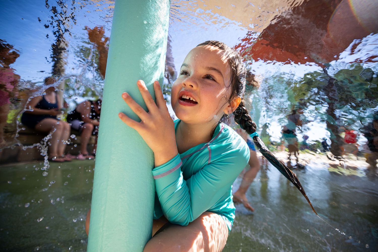 Young girl in teal swimsuit playing at outdoor splash pad water park on a sunny summer day.