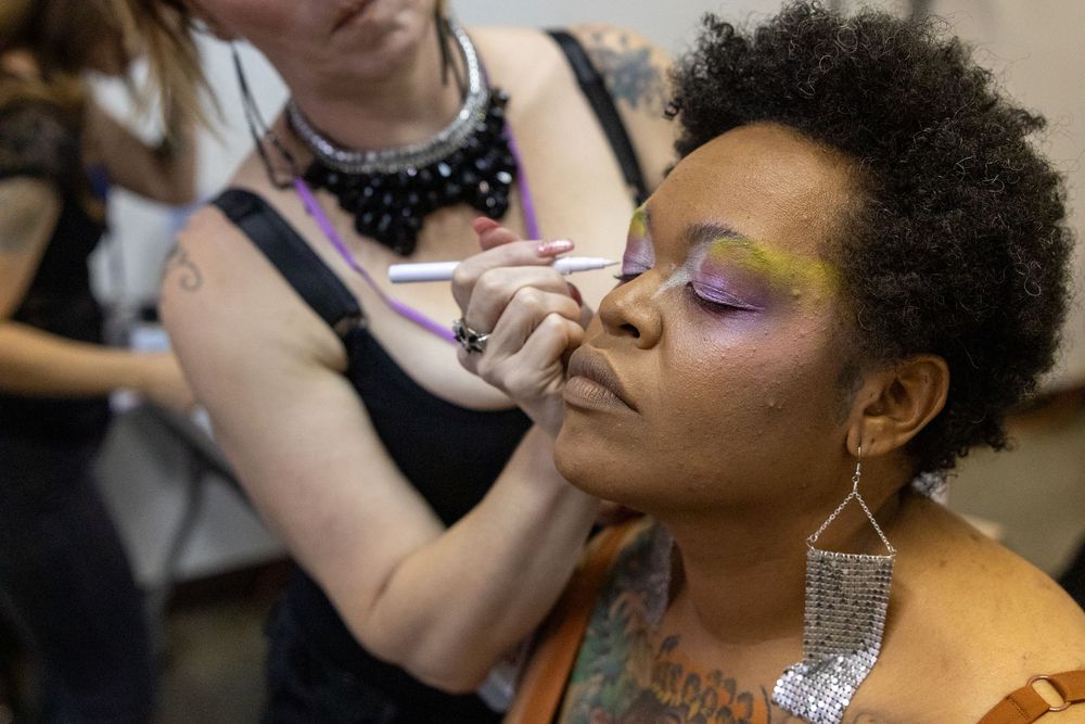 Makeup artist applying colorful eyeshadow to a woman with natural hair wearing silver earrings at a beauty event.