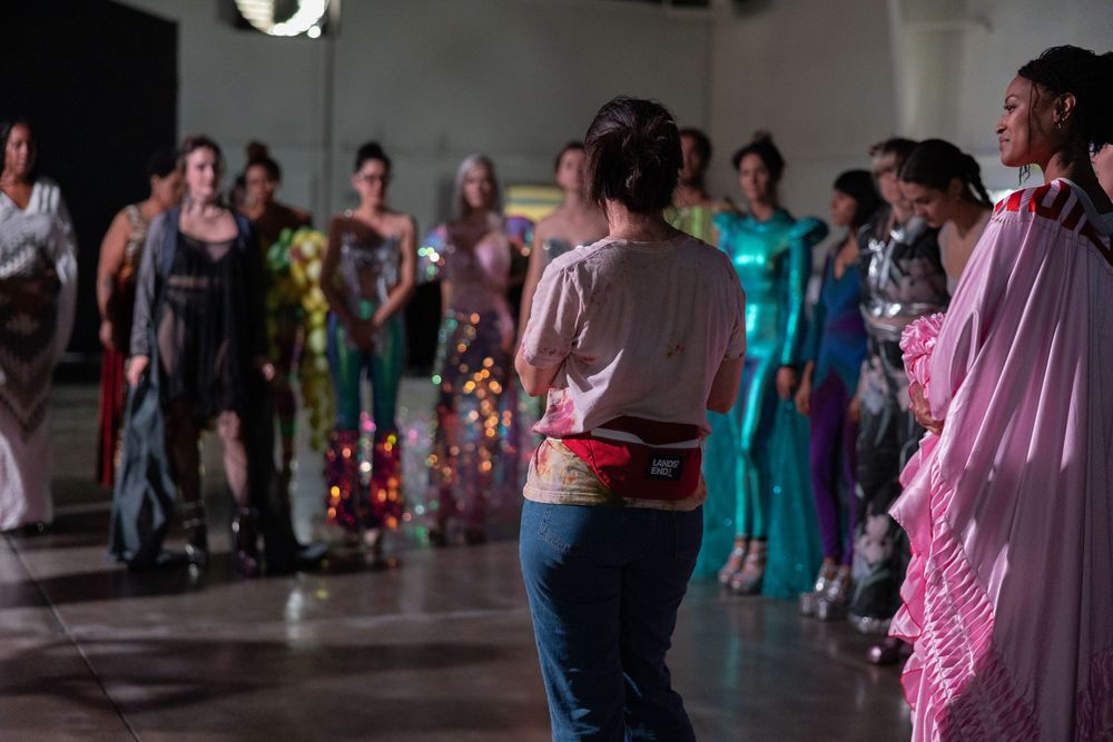 Models in colorful gowns line up at a fashion show while a woman in jeans and a fanny pack observes the runway.