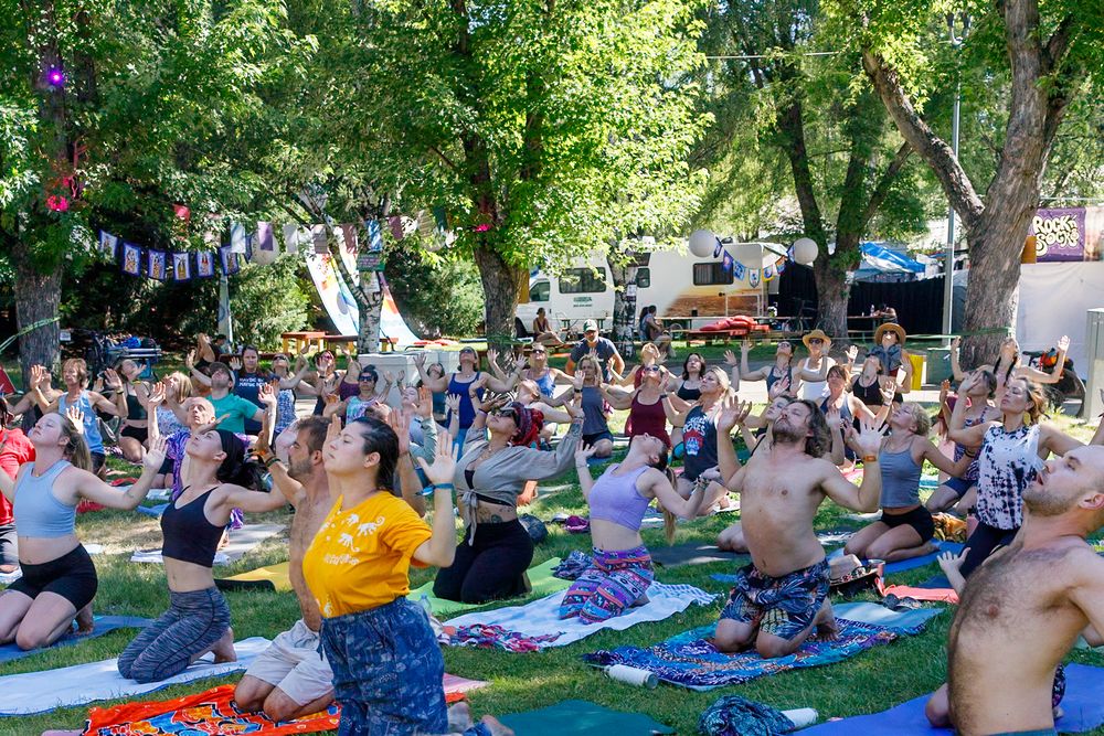 Large group of people practicing yoga outdoors at a festival under shady trees on colorful mats on a sunny day.