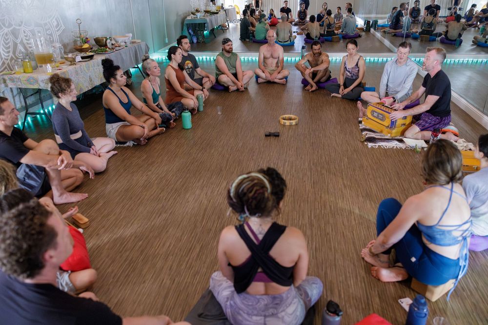 Group of yoga participants sitting in a circle on wooden floor during a meditation or wellness class session.