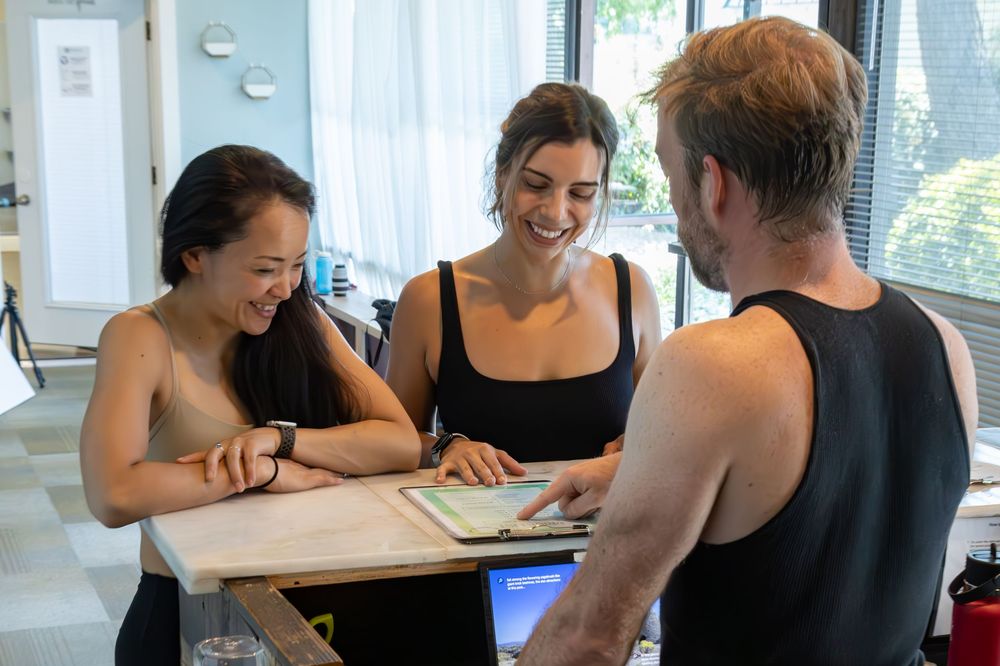 Three people in black outfits smile while reviewing documents together at a bright, modern workspace.