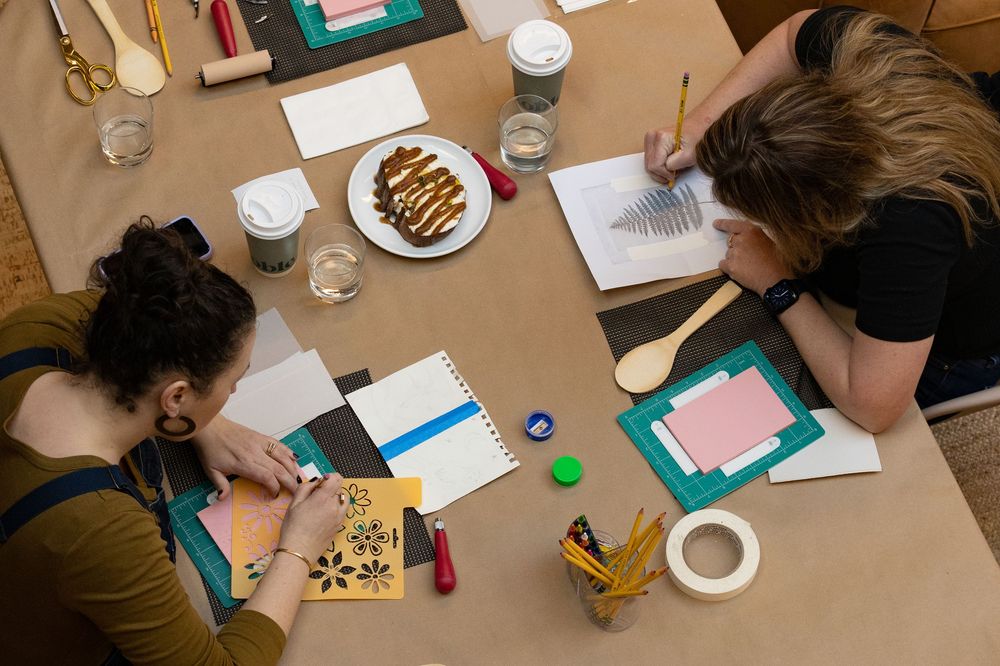 Two women work on creative art projects at a craft table with papers, pencils, and supplies.
