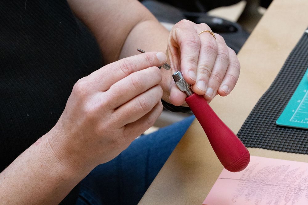 Close-up of hands using a red-handled tool to work on a small object at a crafting table.