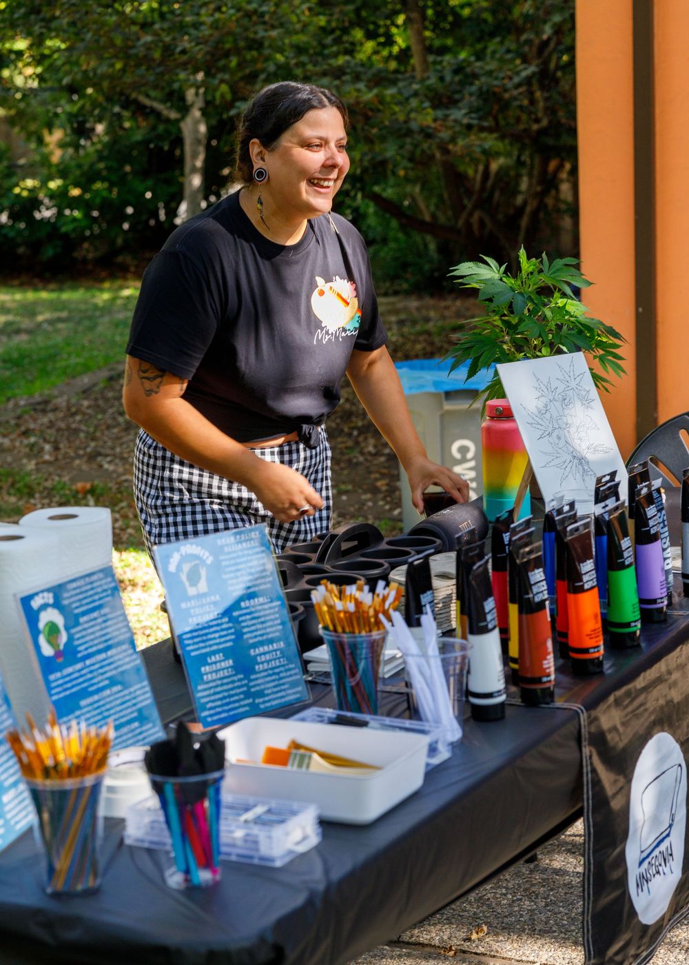 Smiling woman at outdoor art supply booth with colorful markers, brushes, and pencils on a black table.