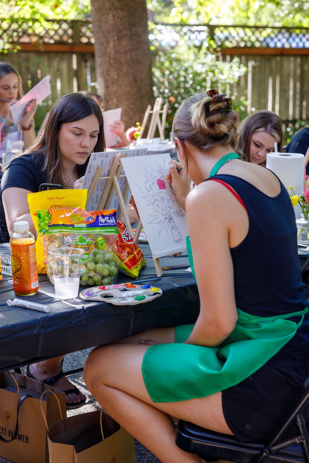 Women painting on canvases at an outdoor art class, with snacks and supplies on a black table in a garden setting.