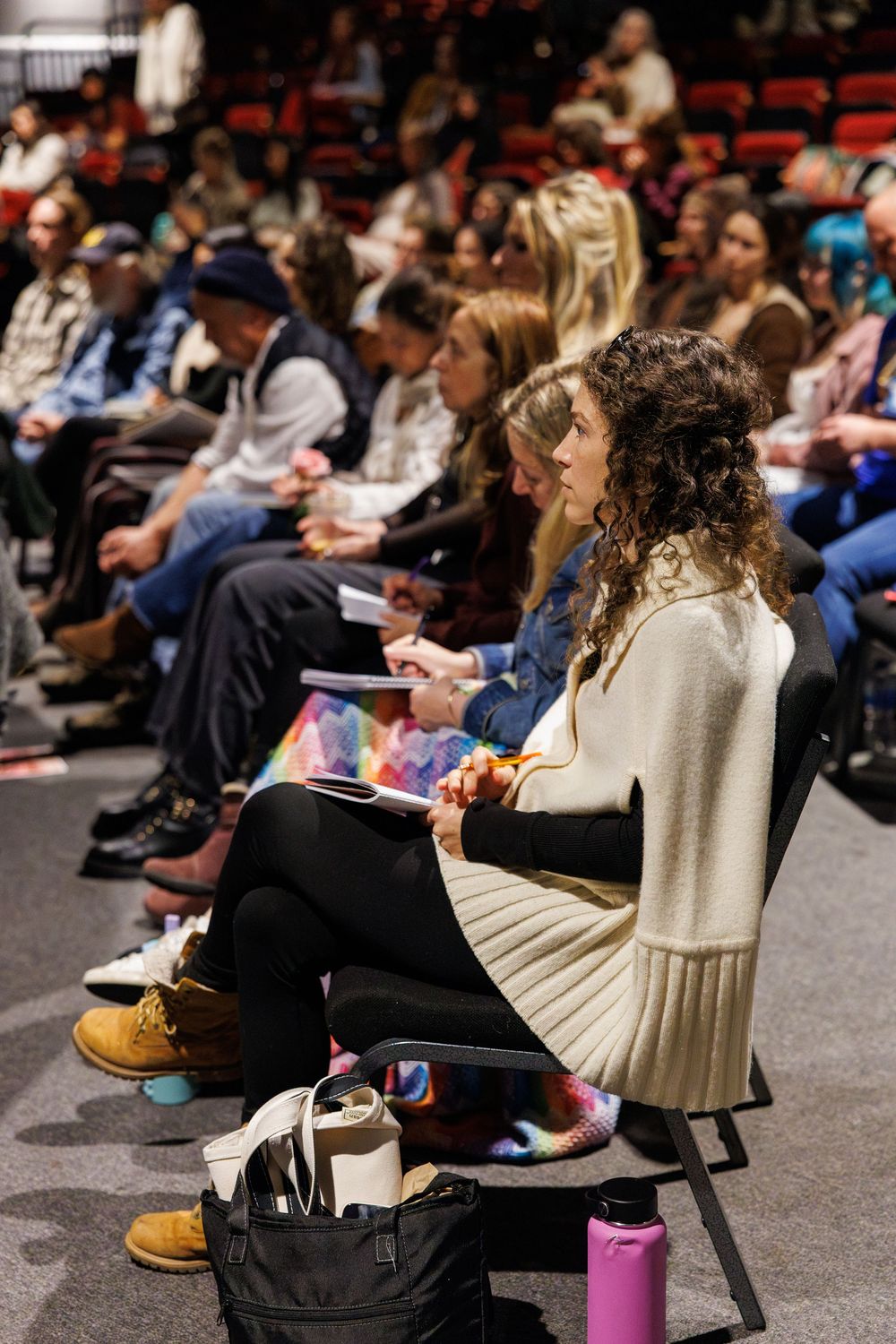 Audience members seated in rows at an indoor event, with a curly-haired woman in a cream sweater taking notes in the foreground.