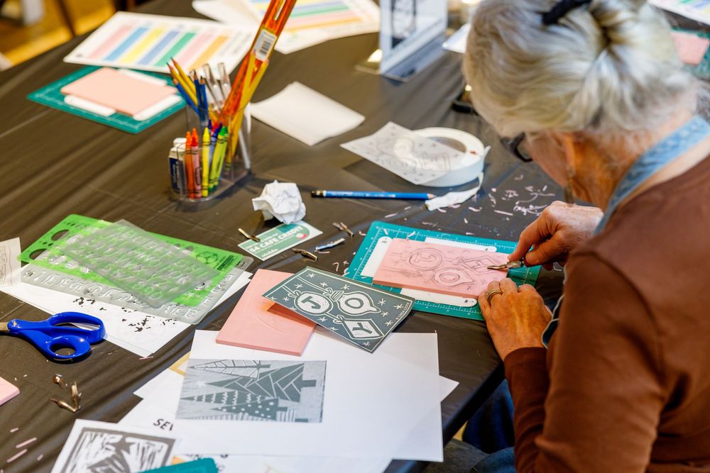 Elderly woman crafting art projects at a table with colorful pencils, scissors, and paper cutouts in a workshop.