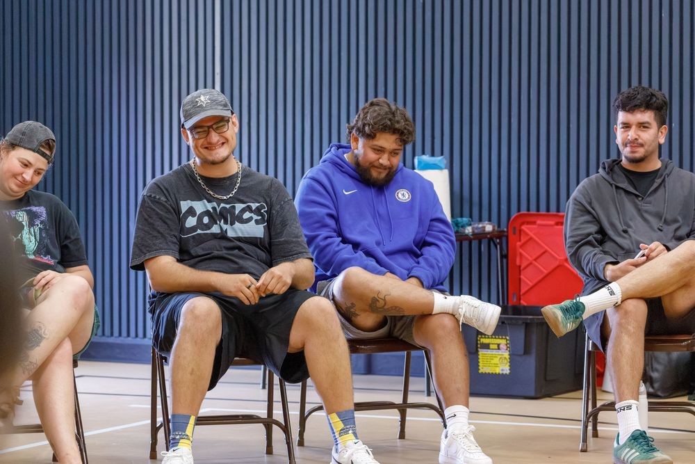 Four young men sitting on chairs in a gym, casually dressed, relaxed and smiling during a group meeting.