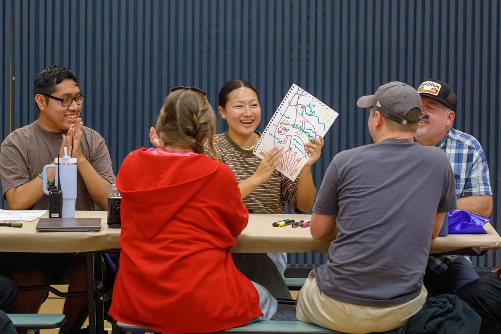 Group of five people sitting around a table, smiling and sharing a colorful drawing during a collaborative activity.
