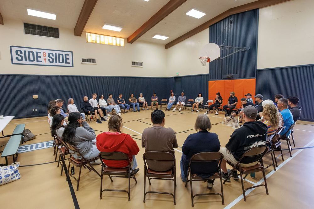 Large group of people sitting in a circle on chairs in a gymnasium with a Side by Side banner on the wall.