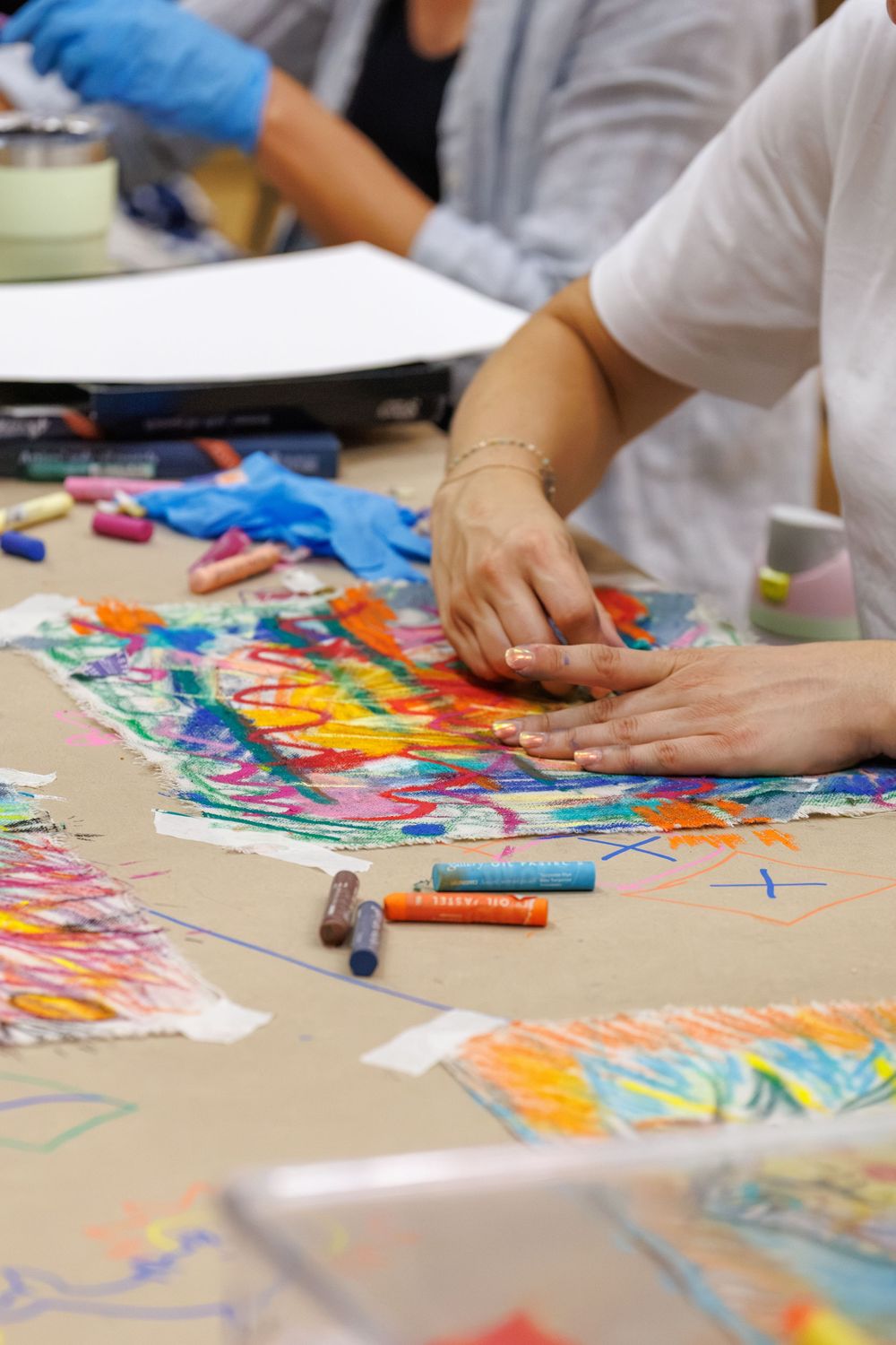 Hands working with colorful oil pastels on vibrant artwork spread across a table in an art class setting.